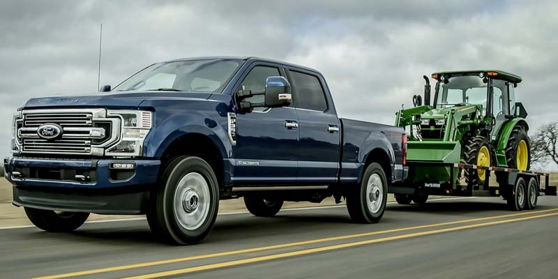 A blue Ford F-250 pickup truck tows a green John Deere tractor on a flatbed trailer on a highway.