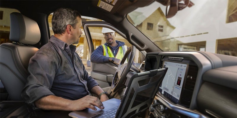 A man on a laptop in a truck's driver seat talks to a construction worker in a hard hat and safety vest.