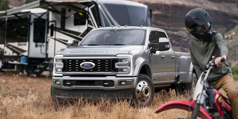 A silver Ford Super Duty truck parked near a large RV. A person on a dirt bike is in the foreground.